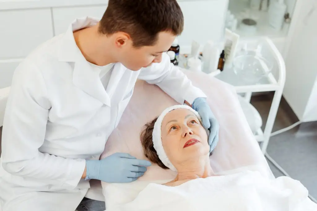Senior, mature woman lying in cosmetic room, cosmetologist doctor doing cosmetic procedures on skin