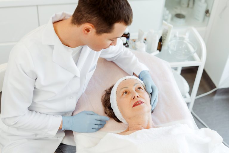 Senior, mature woman lying in cosmetic room, cosmetologist doctor doing cosmetic procedures on skin